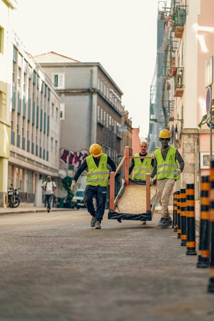 pexels-photo-19250066-19250066 Three construction workers in safety vests transport heavy equipment on an urban street.